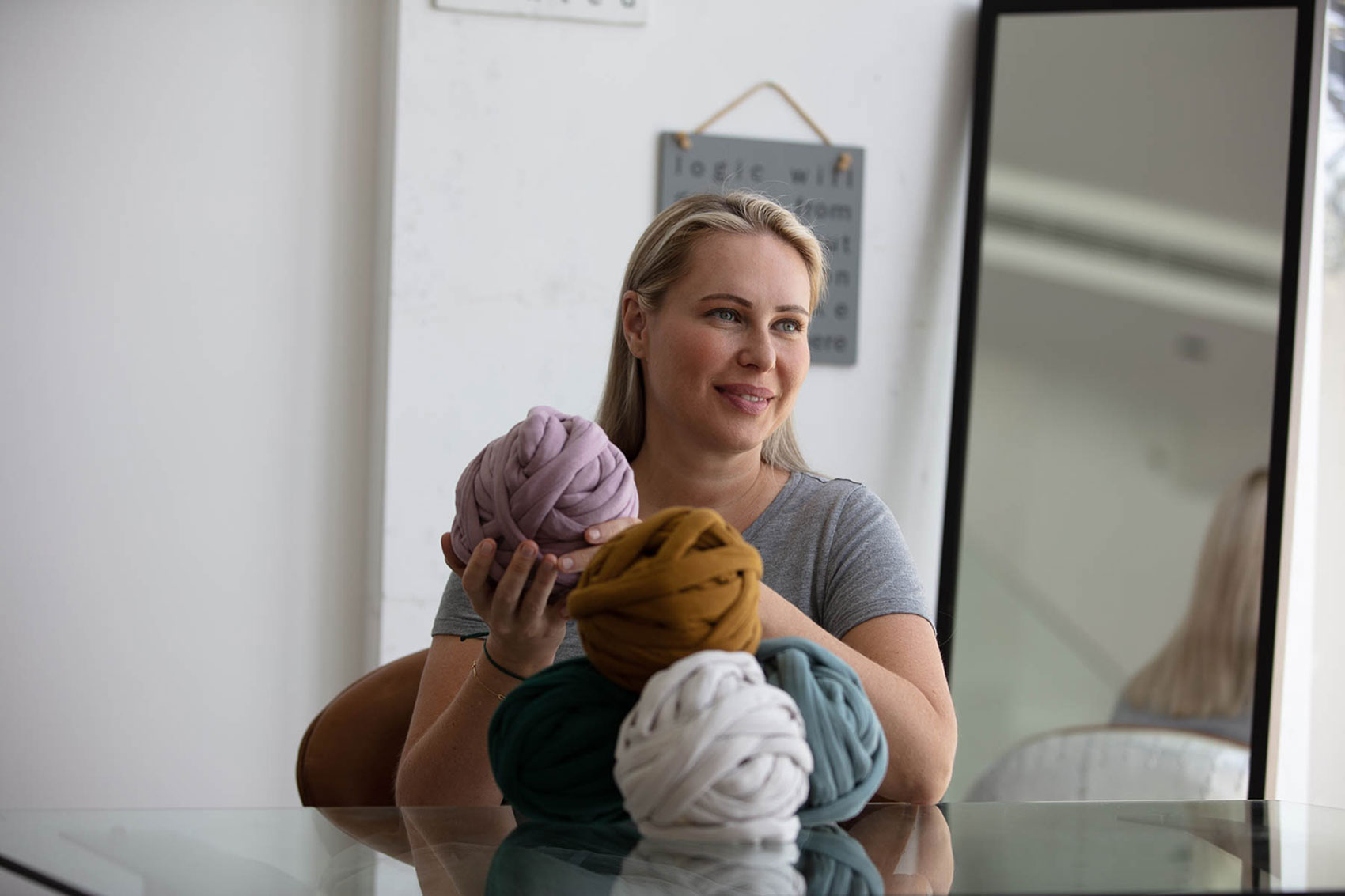 Horizontal image of a blonde-haired woman in a gray t-shirt sitting down at a table and looking to the side of the camera while holding a large ball of light pink/lavender yarn in her hands. Resting on the table in front of her are four other balls of yarn stacked in a pyramid with a mustard yellow one on top, a dark turquoise one to the left, an off-white one in the middle and a blue/gray one to the right. A white wall sits in the background with a gray, square painting hanging on it and a large full-length mirror resting to the side. 