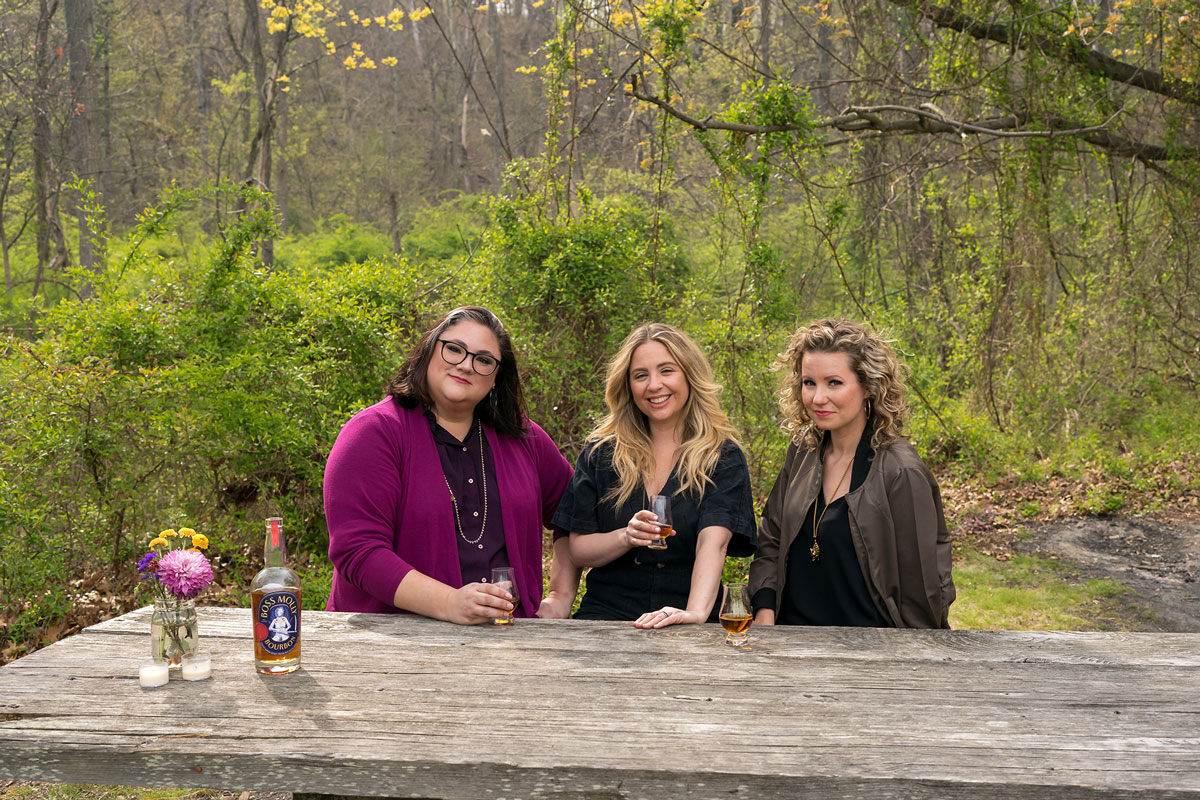 Horizontal image of three women sitting at a rustic wooden picnic table outside in a wooded green area. A bouquet of fuschia, purple and yellow flowers are sitting in a mason jar on the table to the left with two small white tea lights and a bottle of amber-colored bourbon. The woman to the left has short dark hair, glasses, and is wearing a purple and fuschia outfit. The woman in the center has long dirty blonde hair and is smiling widely with a glass of amber bourbon in her hand. The woman to the right has a glass of bourbon resting on the table in front of her. She is wearing an army green bomber jacket and has curly, shoulder-length dirty blonde hair. 