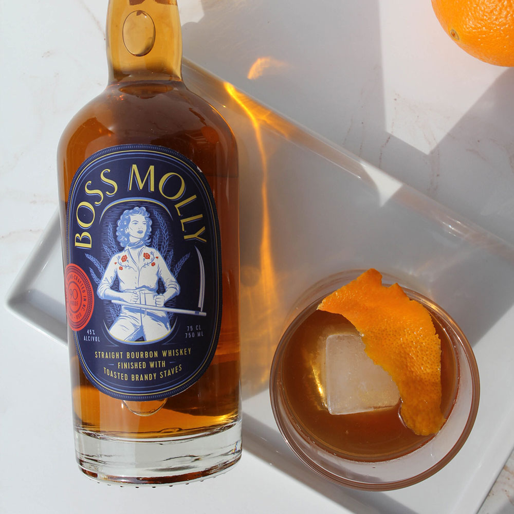 A close up, circular, product image taken from an aerial view of a bottle of amber bourbon to the left and a a glass filled with an amber cocktails and ice cube and topped with an orange peel. The label on the bourbon bottle is dark blue with light yellow lettering. In the center of the label is a woman sketched in light blue and white dressed in a collared shirt with dark pink flowers, pants, and a neck scarf. She is holding a large scythe. There is a scarlet colored circular seal to the left of the woman. The background of the image is white marble tiles. 