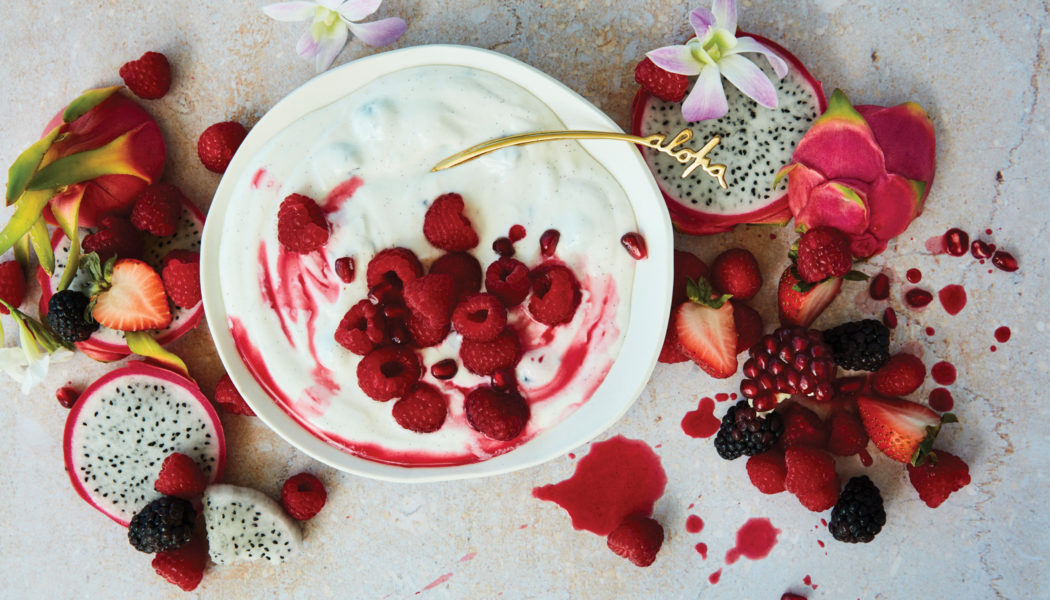 Bowl of yogurt topped with raspberries and streaked with puree. The table is covered in fresh fruit; raspberries, black berries, strawberries, and dragon fruit.