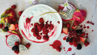 Bowl of yogurt topped with raspberries and streaked with puree. The table is covered in fresh fruit; raspberries, black berries, strawberries, and dragon fruit.