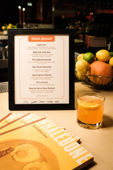 A close up image of a white cocktail menu standing up in a black picture frame on an off-white table. A bowl of oranges, pink grapefruits, green limes, and yellow lemons is in the background behind the menu to the right. In front go the menu to the right is a small whiskey glass filled with an orange drink and topped with a lemon wedge. In the foreground to the left, the tops of three Whalebone magazine covers are fanned out.