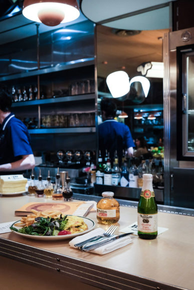 Image of a full breakfast setting on a diner bar. Resting on the bar are utensils wrapped in a napkin, a plate full of eggs, fries, and mixed greens and a small roundd bottle of golden Martinelli's apple juice. Next to the small bottle is a taller green bottle of Martinelli's apple juice with a foil-wrapped neck. Behind the bar is a man in a blue shirt preparing drinks and a full bar with a mirror and three shelves of bottles.
