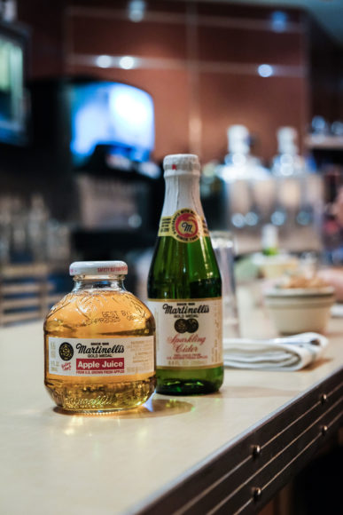 Close-up, focused image of a small rounded bottle full of golden Martinelli's apple juice in front of a tall green bottle of Martinelli's apple juice with a foil wrapped neck. Both bottles are resting on a diner bar with napkins and coffee ups behind them as well as the back of the bar visible in the background.