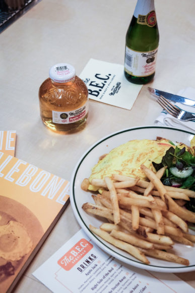 Semi-aerial image of a breakfast meal with orange whalebone magazines scattered to the left, a heaping plate full of fries, eggs, and mixed greens, a menu to the left of the plate, forks and knives setting the right of the plate. a short rounded bottle of Martinelli's apple juice in front of the plate to the right and a tall green bottle of Martinelli's apple juice resting on the edge of a napkin.