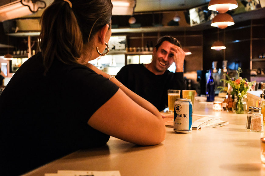 A close up image of a man and a woman leaning on a diner bar and talking. The woman's back is facing the camera and the man is smiling with his hand resting on his forehead and his elbow resting on the bar. A full glass of light yellow liquid sits in front go the man and a white and blue can of beer sits in front of the woman.