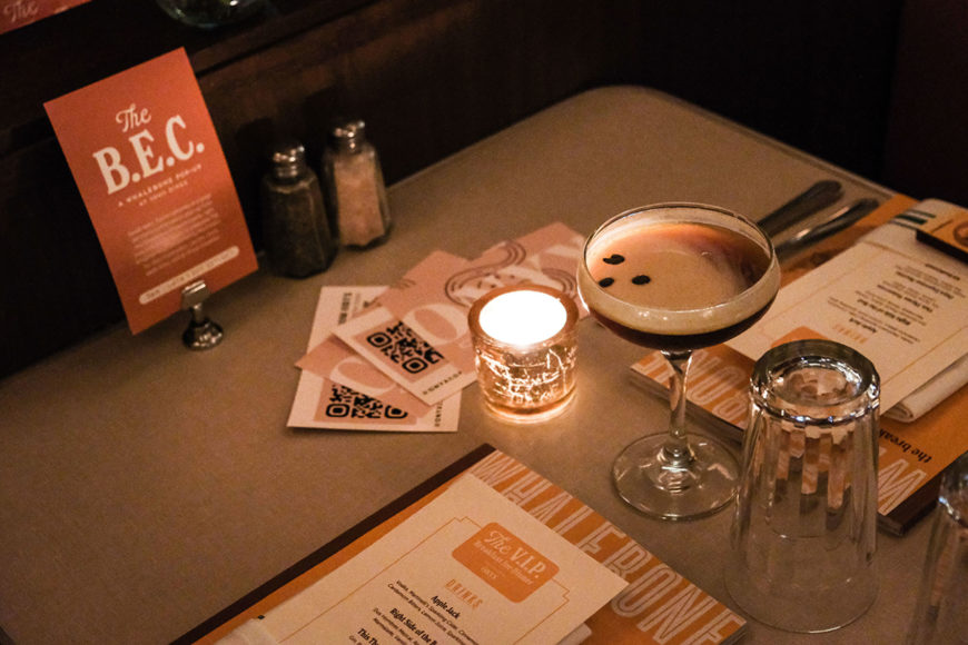 A dimly little close-up image of a table setting. The table is set with a small glass tea light, salt and pepper, and a small orange specials card. Each place setting includes an orange Whalebone magazine with a rectangular white menu on top. In between the two place settings is a shallow, wide-rimmed martini glass filled with a dark brown beverage and topped with a layer of beige foam. next to the glass is a small yellow tea light and an empty glass turned upside down.