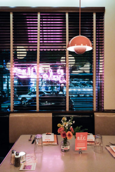 An image taken from just above table level of a diner booth with a window in the background. On the outside of the window is a light pink/purple LED sign that says "Soho Diner" in cursive letters. The sign is also little by an electric aqua blue LED light on the bottom ,giving and ombre effect of purple to blue. The inside of the window is covered with blinds. The table and chairs are an off-white/beige and the table is set for two with a small bouquet of yellow and purple flowers in the center.