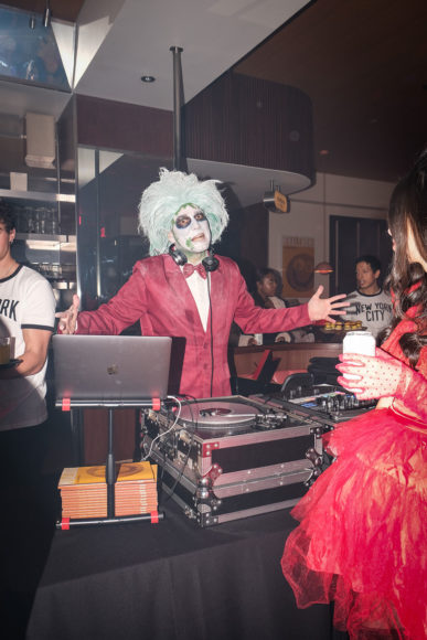 An image of a DJ with a black sounds board and a grey Mac computer in front of him. The DJ is dressed up as Beetlejuice and is wearing a light aqua long-haired wig, white and black face paint, and a bright red suit. The man is holding his hands out in presentation. In the bottom right corner, a girl in a bright feather red dress holding a white beer can is partially visible.