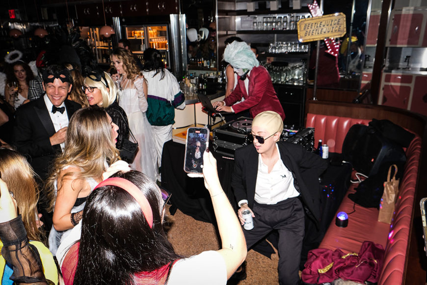 An image taken with flash of crowd is dancing around a person dancing in the corner while wearing a black suit, a bald cap, and sunglasses and holding a drink. The woman closet to the camera is holding up her phone to talk e a video. She is wearing a red dress and a red headband. A DJ in the backgrounds wearing a light aqua, long-haired wig and a red suit while playing music on the sound black sound deck in front of him.