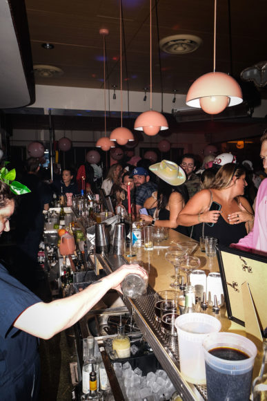 An image taken with flash from the end of the bar. The backside of the bar is filled with alcohol bottles and a bartender is in the middle of pouring a drink. The bar is lit by a line of pink lamps hanging from the ceiling in a straight line. A crowd of people are gathered on the frontside of the bar.