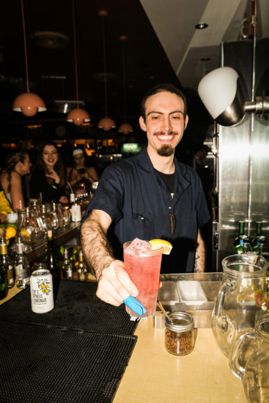 A portrait image taken with flash of a male bartender with a dark goatee and mustache smiling as he hands the photographer a large glass filled with a salmon-colored drink and topped with a lemon wedge across the bar. The bartender is wearing a black button down shirt and has a light blue badge on his finger. The bar is covered with glasses and beer cans. In the background, behind the bar is lined with lots of alcohol bottles. People are sitting on the other side of the bar in the dimly lit background.
