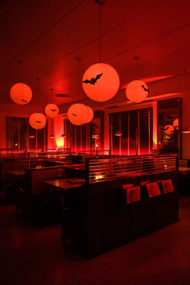 An image taken of a diner lit lowly by a harsh red light. Large red orbs hang from the sealing as lights along with fake bats. Windows lines the walls and the booths are setup in a grid-like fashion. Three Whalebone magazines hang from the back of the closest booth.