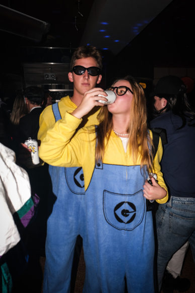 A close-up image taken with flash of a man and a woman dressed up as minions. They are both wearing marigold yellow, long-sleeve shirts with denim blue overalls that have a black symbol on the front. The man is standing behind the woman and wearing black sunglasses. The woman is wearing black framed glasses and standing in front of the man while drinking deeply from her white beer can.