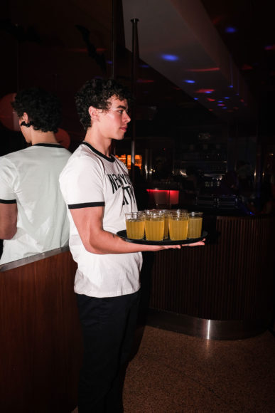 And image taken with flash of a male server's profile. The server is standing tall and straight while wearing a white t shirt with black writing and black lined sleeves. The back of the man is visible in the mirrored wall that he is standing against. The man is holding a tray full of full glasses with golden yellow drinks. The background is very dark with a red hue.