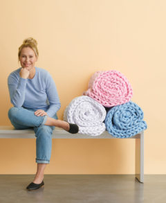 A feature image of a blonde-haired woman in a light blue t-shirt, jeans and black ballet flats sits on a long white bench against a soft, light yellow wall. The woman is smiling with her legs crossed, her elbow leaning on one leg and her chin leaning on a fist as she smiles at the camera. To the right of the woman are three large knit blankets, rolled into spiral shapes and sitting on top of each other in in a pyramid form. One is white, one is baby blue and the top blanket is light baby pink. The cement floor is dark gray.
