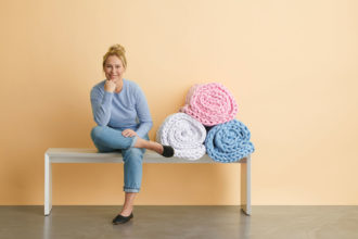 A feature image of a blonde-haired woman in a light blue t-shirt, jeans and black ballet flats sits on a long white bench against a soft, light yellow wall. The woman is smiling with her legs crossed, her elbow leaning on one leg and her chin leaning on a fist as she smiles at the camera. To the right of the woman are three large knit blankets, rolled into spiral shapes and sitting on top of each other in in a pyramid form. One is white, one is baby blue and the top blanket is light baby pink. The cement floor is dark gray.