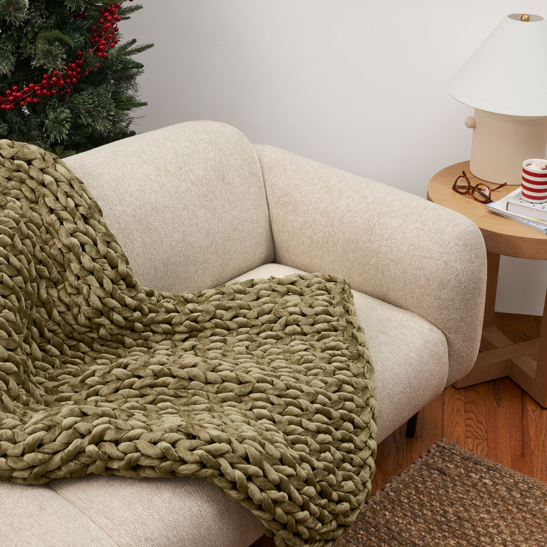 A circular image taken at a horizontal angle of a couch with light beige/off-white cushions and a large olive green, woven weighted blanket draped over the front and back. At the bottom of the image, part of a medium brown woven rug is visible. Next to the couch, on the right side of the image is a small, light brown, circular wooden side table. A pair of red glasses, a stack of books, a red and white striped mug of brown liquid topped with white cream and a light beige lamp with a white lampshade resting on the table. Behind the couch, the dark green branches of a pine tree decorated with bright red holly berries is visible. 
