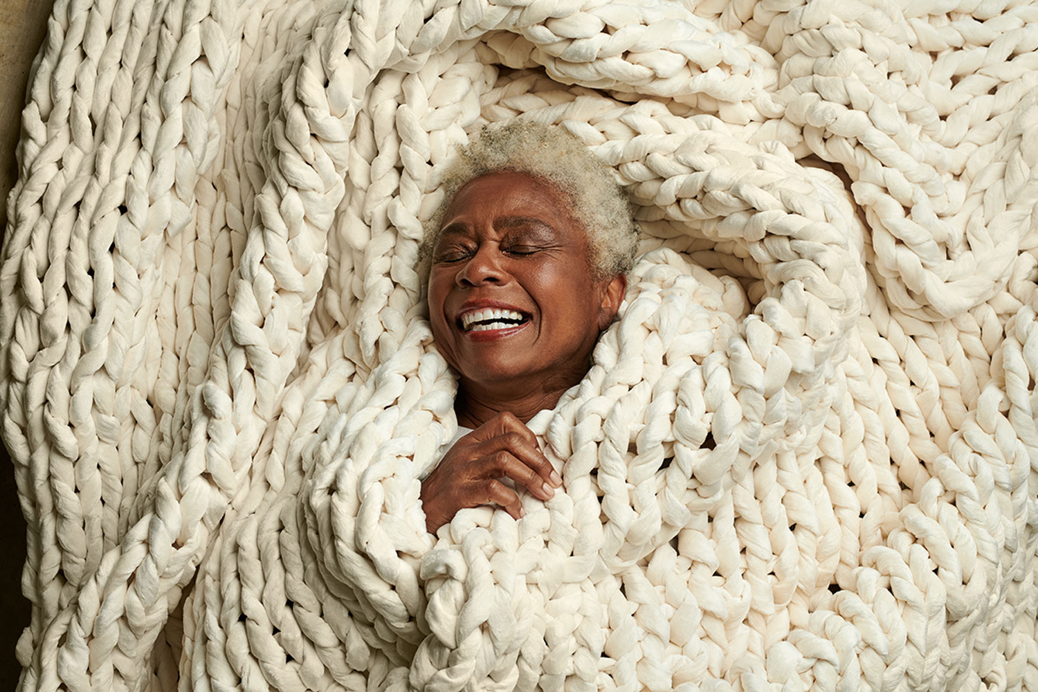 Aerial image of a gray-haired woman laying down and wrapped up in a large, off-white knit throw blanket. Only her face and one hand is visible. She is smiling widely with her eyes closed in a joyful manner.