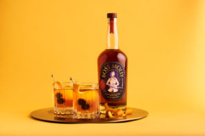 A horizontal feature image with a completely sunflower yellow background. Just off-center is a round, mirrored serving dish with two glasses filled with light orange cocktails, each with two blackberries and an orange peel slice skewered on metal stir sticks. To the right of the two glasses on the serving dish is a bottle of unopened amber bourbon with a navy blue label that has gold text and the white silhouette with red accents of a woman holding a ranching scythe.