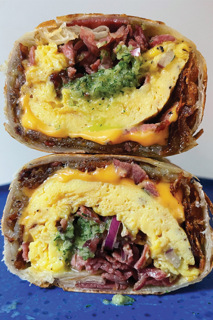 A breakfast burrito that has been cut on and each half stacked on top of one another so you can see all the filling inside. The burrito is filled with egg, pastrami, cheese, pickles, potatoes, and onions.