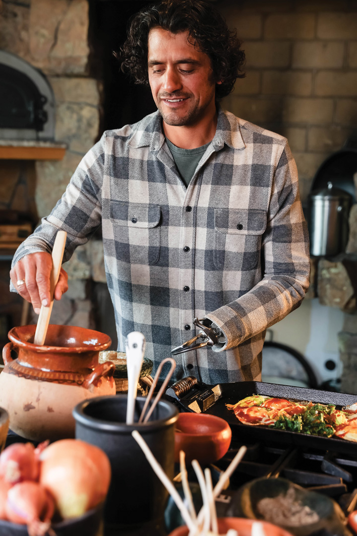 Eduardo Garcia stirring a pot near the stove. The pot is more of a vase shape and is a terracotta color. The kitchen counter is filled with ingredients, utensils, and something cooking on a cast iron skillet on the stove. 