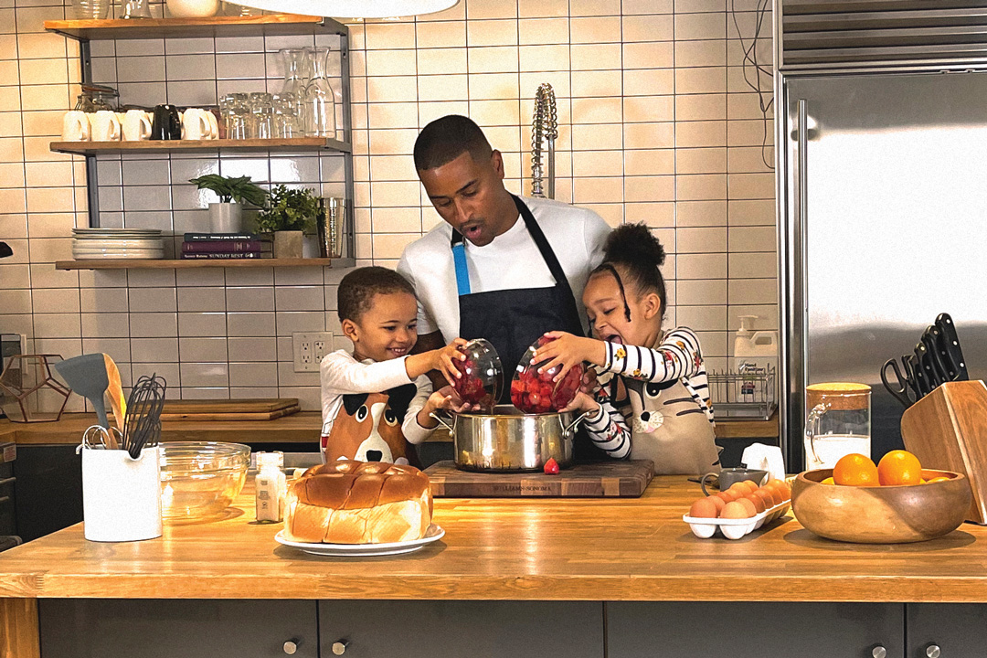 JJ Johnson cooking in the kitchen with two young kids. The kitchen island is filled with ingredients and utensils, and the two kids are pouring bowls of berries into a sauce pan while Johnson watches.