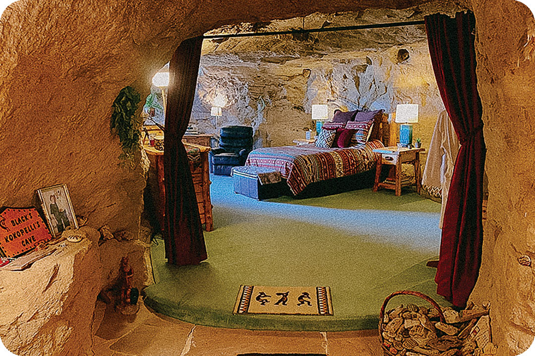 Inside view of a room at Kokopelli's cave. The walls are all carved from rock. There is a small step up into the room, the floor is covered in a green carpet and red curtains are drawn to the side so you can see into the bedroom. Inside the room is a medium-sized bed, a dresser, nightstand, and a reclining chair.