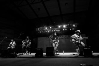 Black and white photo of our men sit on stools on a large stage. They each have a guitar in their hands and a mic sitting in front of them. Behind them are a set of large lights that are illuminating the stage.