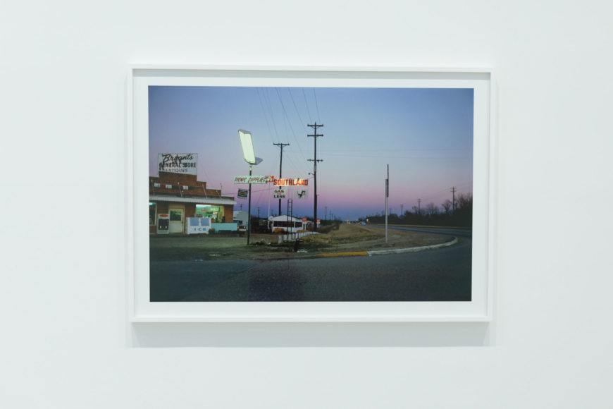 A photo of a horizontal print photograph set in a white frame against a white wall. The framed image is of an old, run-down general store taken from across the street at dusk. There are telephone lines and street lights surrounding the white store building. A few old-fashioned cars are parked in the parking lot. An endless green field lies behind the building and an indigo sky fades to a hazy pink sunset in the background.