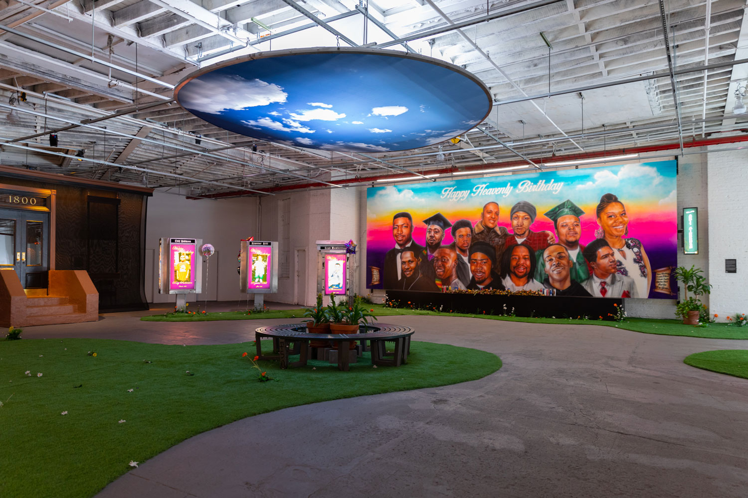 A landscape image of an art exhibition that looks like an indoor park. A large, colorful banner painted with the portraits of 13 people hangs on the back wall. A large circle with a picture of a partly cloudy, blue sky hangs from the ceiling. Gray sidewalks curve around sections of green turf adorned with the occasional sprout of red carnations and scattered dead leaves. Three neon signs are in the background to the left. A small, dark brown circular bench is in the middle of the room with plants in the center of it. 