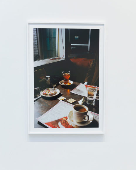 A photo of a vertical print photograph at an art exhibit set in a white frame against a white wall. The framed photo print is a close up photo of an old-fashioned diner table setting. The lighting is dark and the table and walls are a dark brown. There is a window with white curtains that the table itself sits against. The booth seat visible in the center of the image is a dark red. The table is set with red and white menus, a full cup of coffee, condiments, a glass of water and a glass of amber liquid.