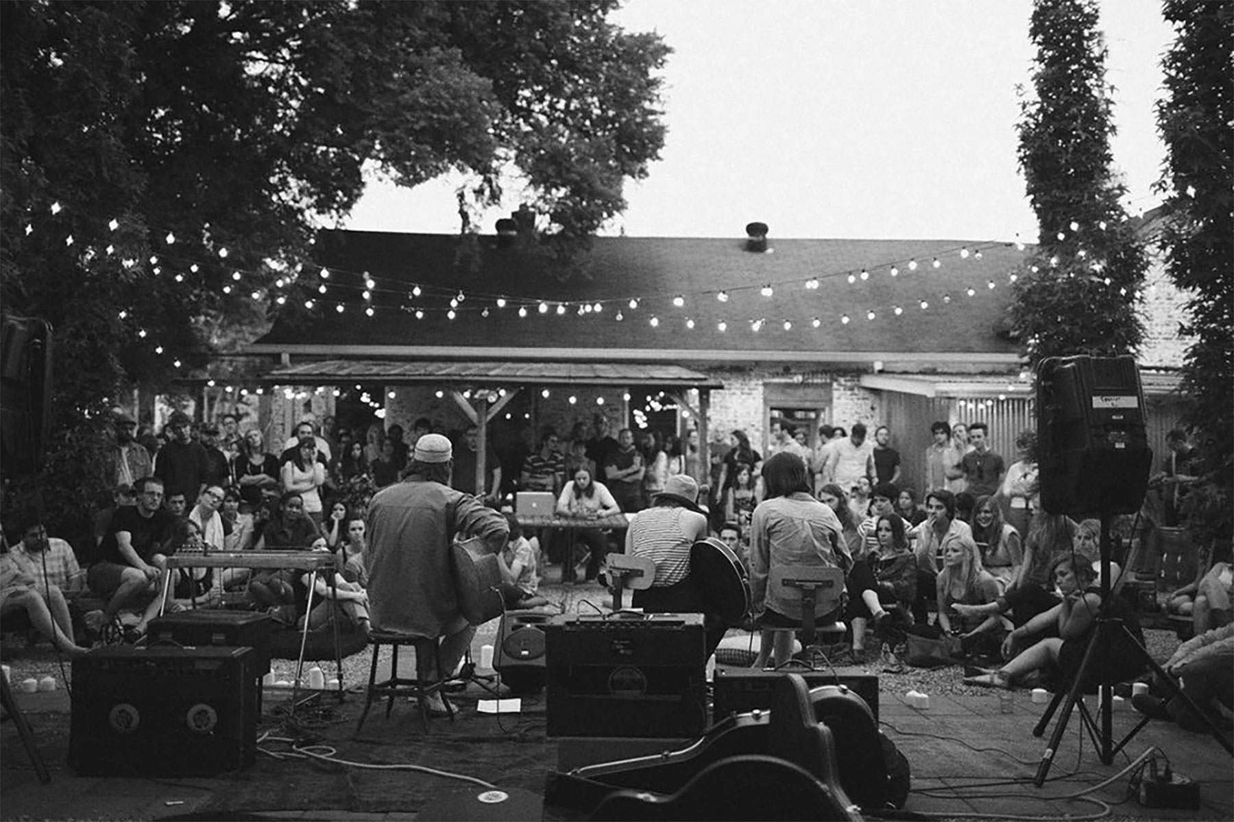 Black and white image of the back of a band sitting casually with their instruments in a backyard. A crowd is watching the band play. There is a brick house in the background, large trees to the left and right, and string lights slung from tree to tree across the image.