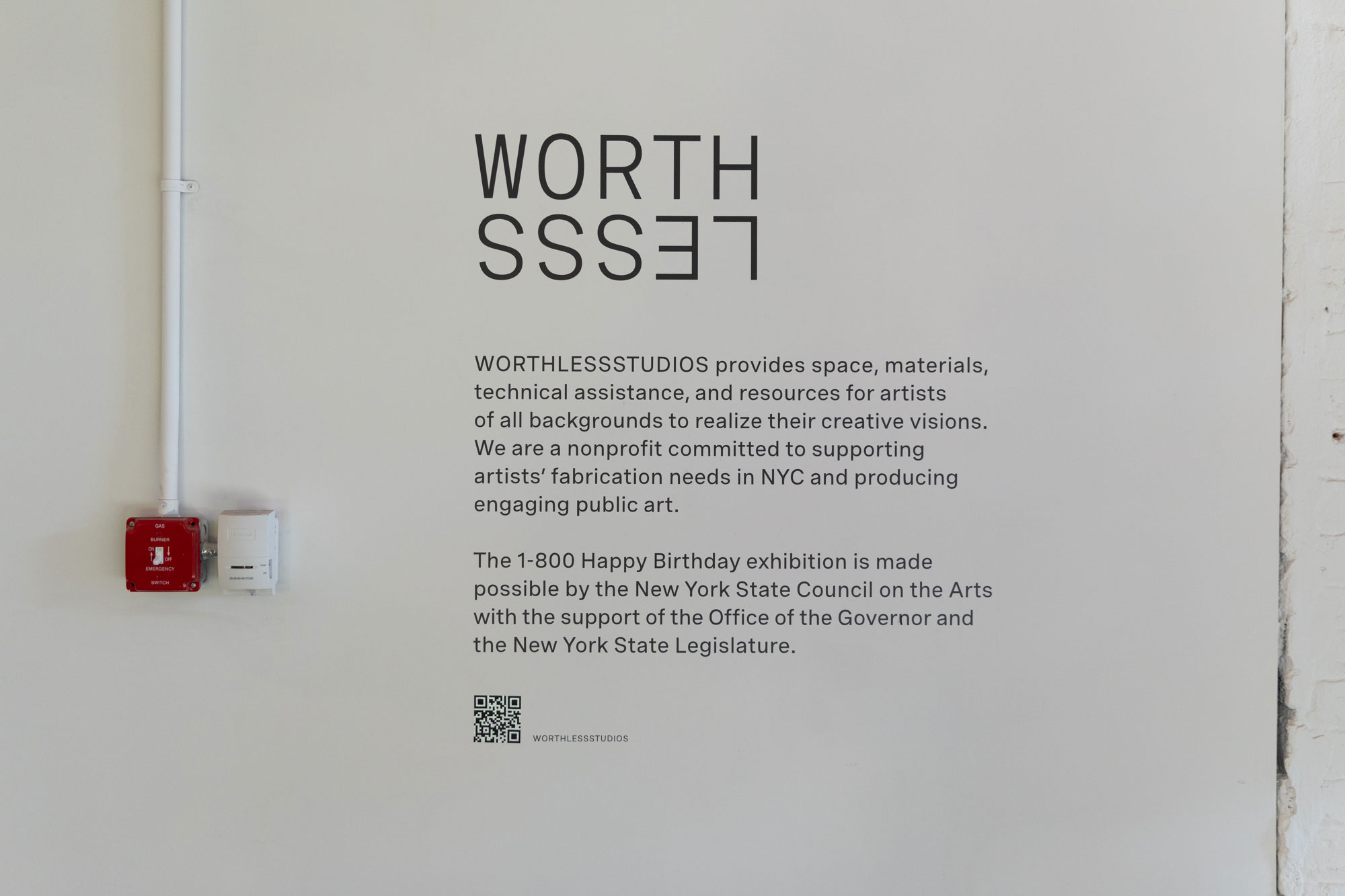 An image of left-aligned black text printed on a white wall. The top text reads "WORTHLESSS" in all capital letter and is larger than the two paragraphs below. It is positioned so that the word WORTH is on top of the second half of the word, LESSS, whose letters are inverted and flipped upside down. A small QR code is positioned underneath the second paragraph. A small, bright red gas burner emergency switch hangs on the wall to the left of the text. 