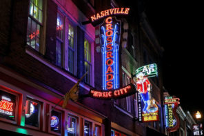 Hero/feature image taken at night from below of the red, green, purple, yellow and blue neon lights of shop and bar signs lining the building of a street in Nashville. At the center of the image is a sign with "Nashville" written horizontally in all caps across the top in red neon lights, the word "crossroads" stacked vertically just below, also in red neon lights and lined with bright blue neon lights and resting on the horizontal, red-lit words "music city".