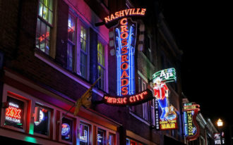 Hero/feature image taken at night from below of the red, green, purple, yellow and blue neon lights of shop and bar signs lining the building of a street in Nashville. At the center of the image is a sign with "Nashville" written horizontally in all caps across the top in red neon lights, the word "crossroads" stacked vertically just below, also in red neon lights and lined with bright blue neon lights and resting on the horizontal, red-lit words "music city".