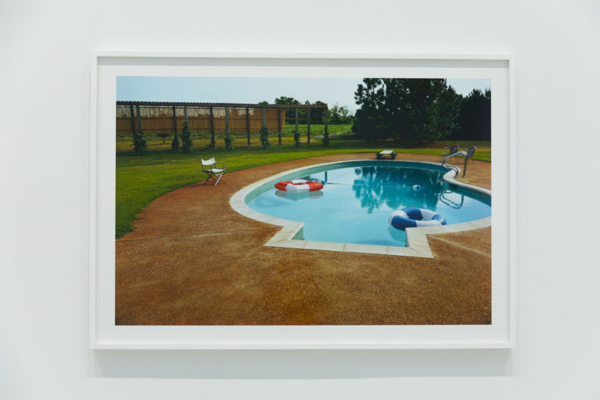 A photo of a horizontal print photograph at an art exhibit set in a white frame against a white wall. The framed photo print is of an old 70s-style in-ground pool filled with light aquamarine water. Dirty white tile lines the curvy perimeter of the pool. Two inner tubes are floating in the water, one red and white striped, the other blue and white striped. Three old lawn chairs are sitting on the light brown concrete that surrounds the pool. The gloomy sky is just visible in the background as is a small clearing of bright green grass that butts up to a large brown wooden fence and a crop field and large trees. Between the crop field and the pool is an old wooden trellis structure covered in ivy.
