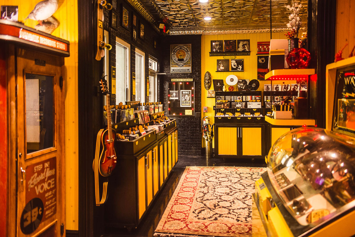 An image of the inside of the Third Man Records shop. The shop is narrow and packed tight with neatly organized records. The wooden walls are painted bright yellow with black accents. The neon red lights of a record stand glows brightly throughout the room. An old, yellow juke box is in the right foreground of the image. Posters line the back, black brick wall that is adjacent to a set of three windows. 