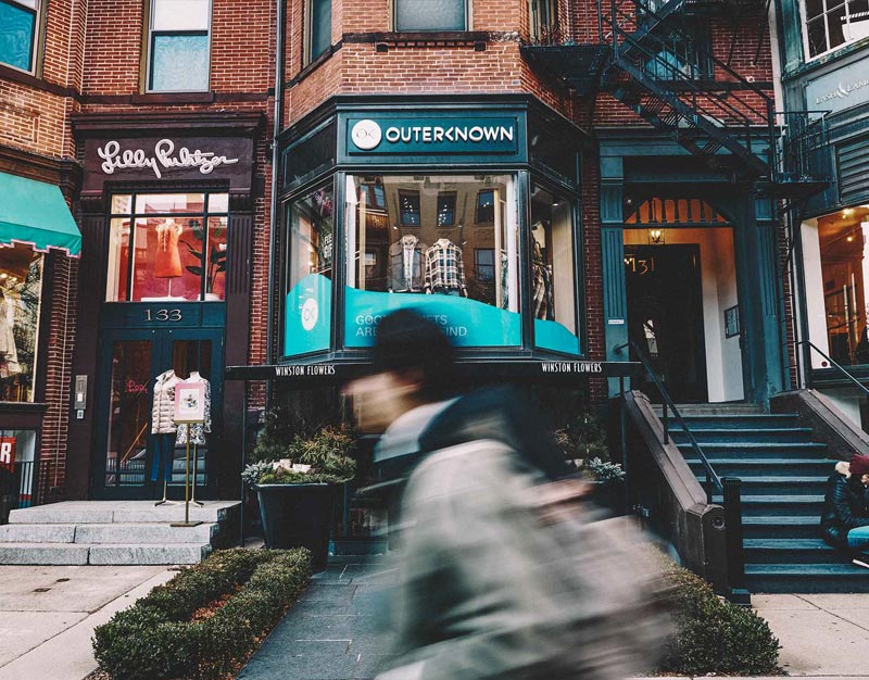 Photograph looking at the front of the Outerknown location in Boston. It's a brick building with the storefront elevated and a large bay window with clothed mannequins on display. In the foreground a blurred person wearing a hat and hoodie walks.