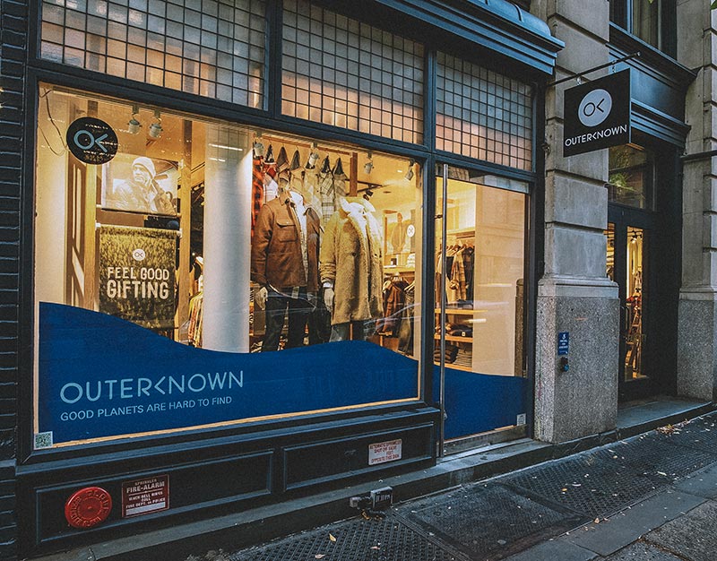 Photograph of the front of the Outerknown store location in Soho. There's a large window and door with clothed mannequins on display.