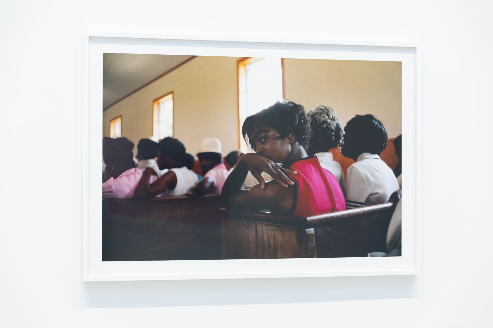 A photo of a horizontal print photograph set in a white frame against a white wall. The framed photo is a close-up image taken from behind of a person in a bright pink dress. The woman is sitting in a crowded church pew and twisted around so that the profile of her face is visible. She has her hand resting on her shoulder and is looking just to the left of the camera. Other people fill the pews in front of her. 
