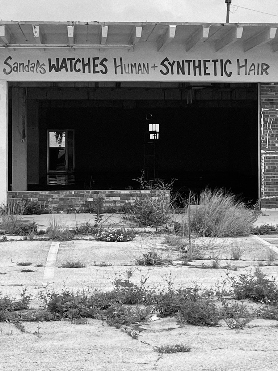 A black and white photo of an old building with brick walls and a white wooden roof with a hand-drawn shop sign. The small building sits in front of a poorly maintained parking lot with weeds growing out of the concrete and cracks everywhere. The front of the building is black and open to the air. A small doorway is visible to the left.