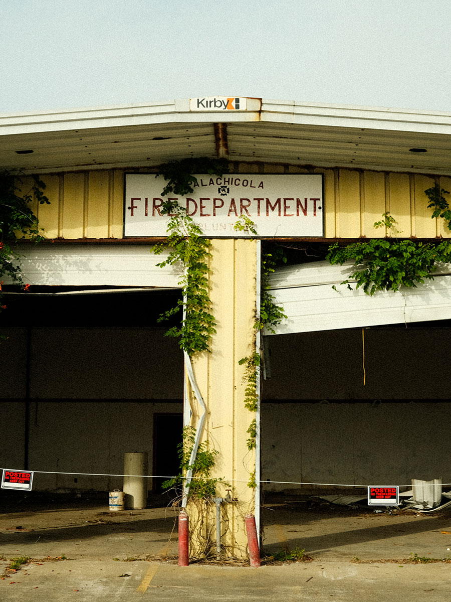 Photo of an old, run down pastel yellow fire department building with vines growing along the front and two open garages with broken white doors side-by-side.