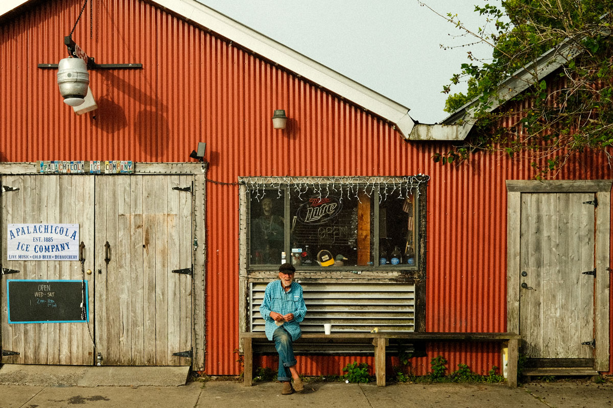 Photo of an old red tin barn with an old man with a short white beard wearing a black hat, blue jeans, and an oversized, bright turquoise plaid shirt sitting on a wooden bench in front of the building with a white styrofoam cup sitting next to him