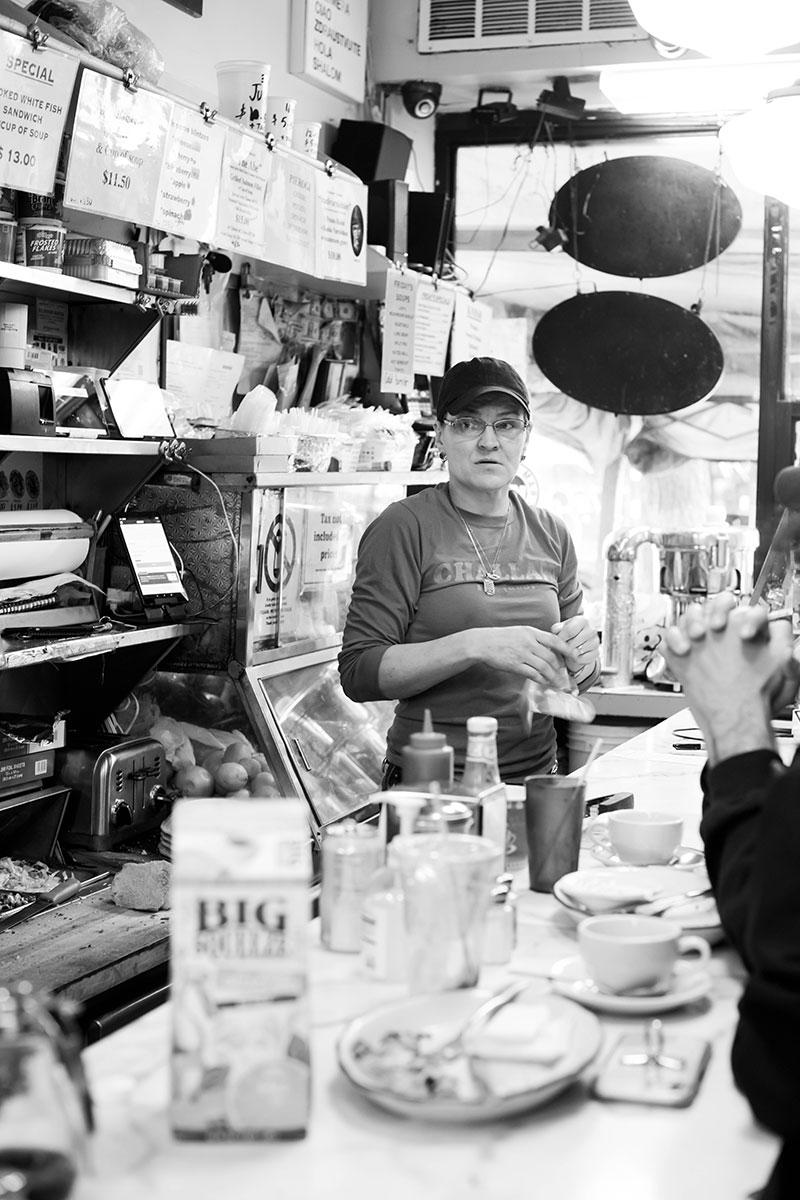 A black and white photo of a woman standing behind a diner counter while serving customers and gazing past the camera. The diner counter is cluttered with dishes and a cartoon of milk.