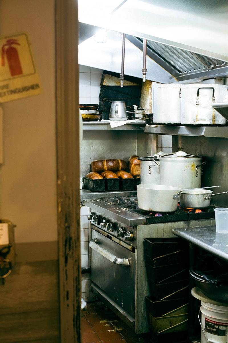 A view of the hidden kitchen of a diner. On the stainless steel, gas stove top are a set up white pots and pans. In the corner on the stainless steel counter, there are stacks of golden brown challah bread loaves.
