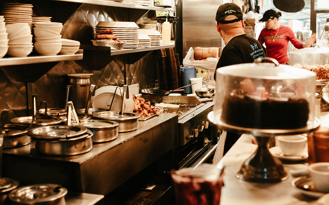 A photo of the backside of a diner counter. The entire cooking/kitchen area is stainless steel and several pots with ladles sticking out of them are lining the back wall. A person in a black shirt is standing in front of the griddle cooking heaps of potatoes and another person in a pink shirt is helping customers. The counter is filled with coffee cups and cake stands with glass lids and chocolate cakes under them. 