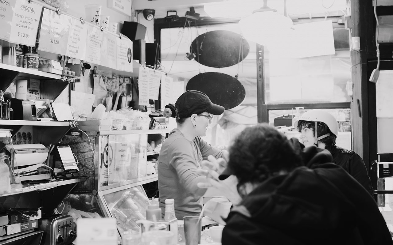 A black and white photo of a woman in a baseball cap with her hair knotted in a bun and her sleeves of her shirt pushed up as she serves customers from behind a diner counter. 
