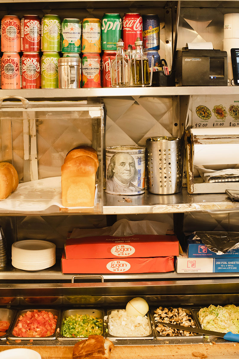 A photo of the back wall of a diner with and array of colorful soft drinks on the top shelf, a roll of U.S. dollars, some notepads and a few loaves of golden bread in a case on the second shelf, red boxes of plastic wrap and blue boxes of aluminum foil on the third shelf down, and a row of stainless steel slots holding various ingredient on the bottom shelf. 