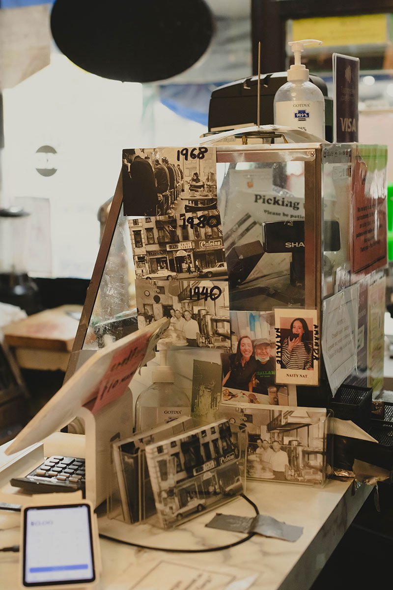 A picture of a cluttered register behind the counter with black and white photos printed through the decades tacked onto the glass display case.x
